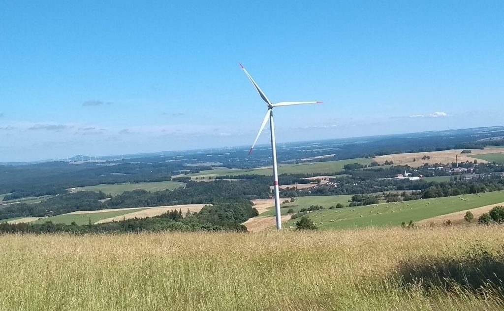 Decentralizované větrné turbíny a farmy by i u nás mohly přispět k přechodu k nízkoemisní elektroenergetice. V popředí osamělá turbína v Jizerských horách, v pozadí nalevo od ní pak skupina turbín v Polsku. (Foto Vladimír Wagner).