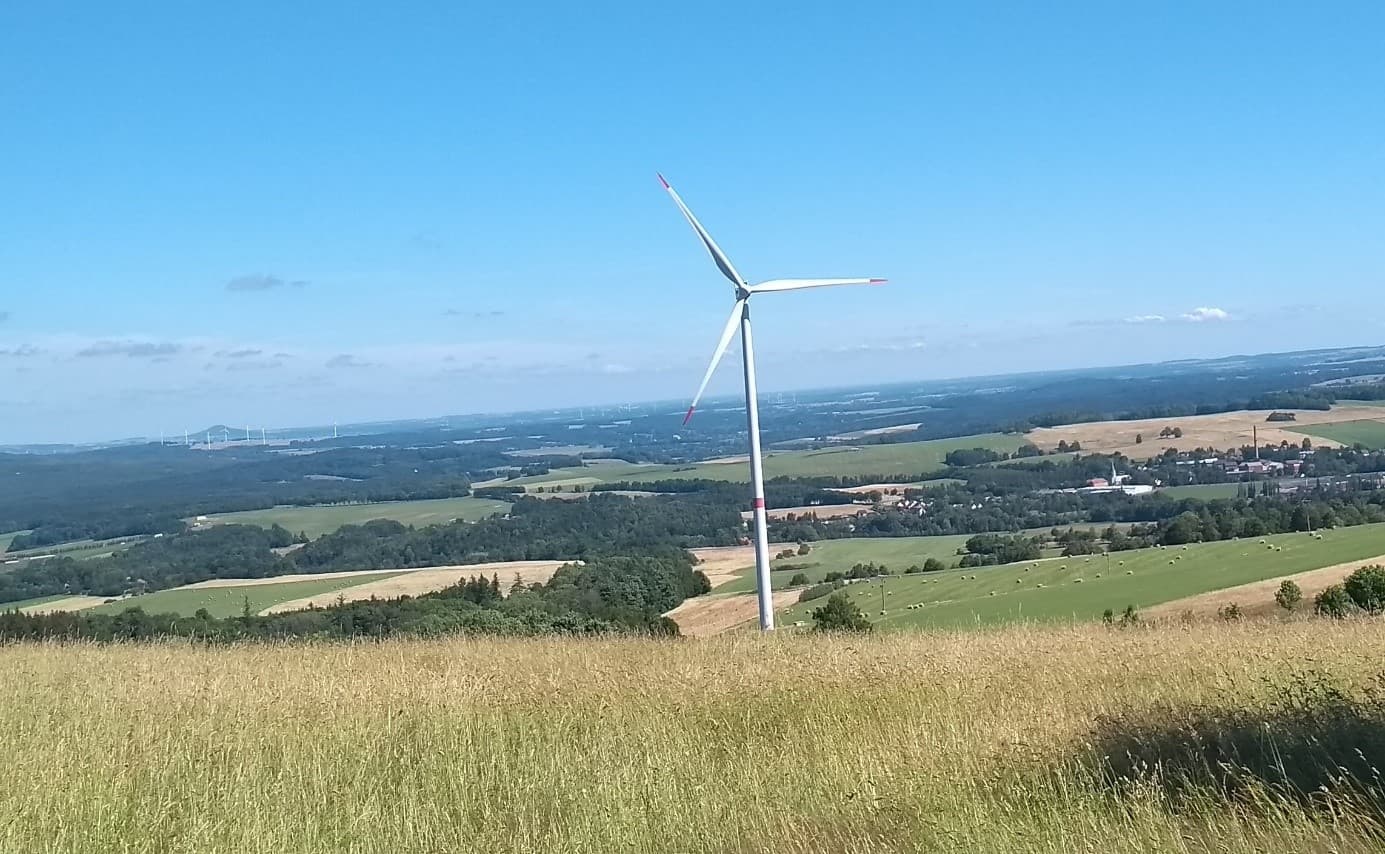Decentralizované větrné turbíny a farmy by i u nás mohly přispět k přechodu k nízkoemisní elektroenergetice. V popředí osamělá turbína v Jizerských horách, v pozadí nalevo od ní pak skupina turbín v Polsku. (Foto Vladimír Wagner).